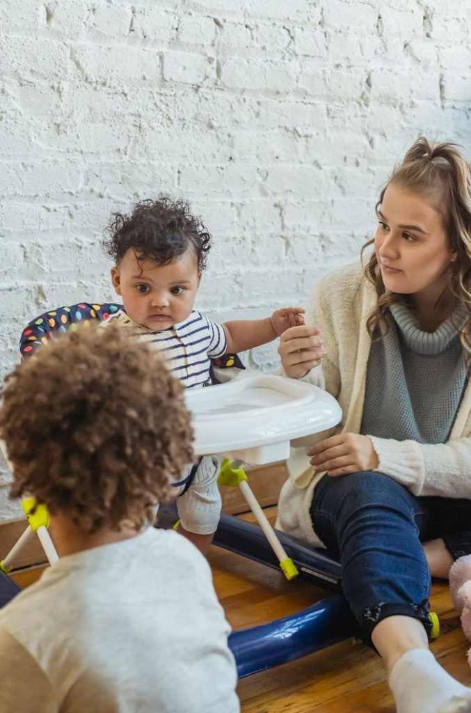 Attentive mother holding hand of little black toddler in baby walker while sitting near wall in room with faceless boy