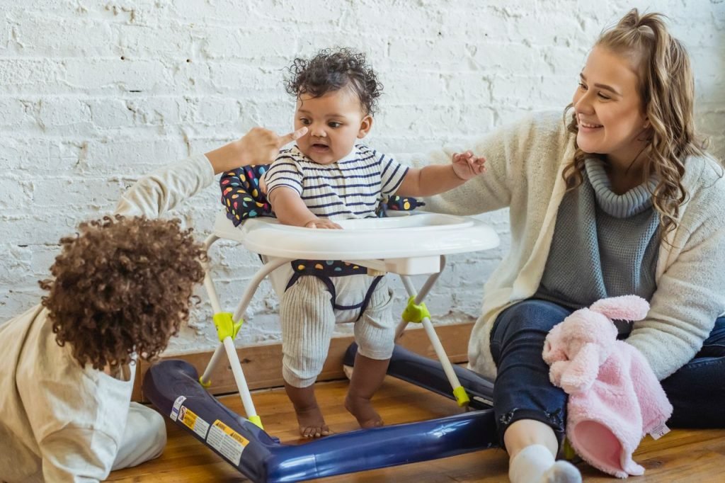 Happy mother with toy playing with black toddler in baby walker and boy while sitting on floor near wall at home