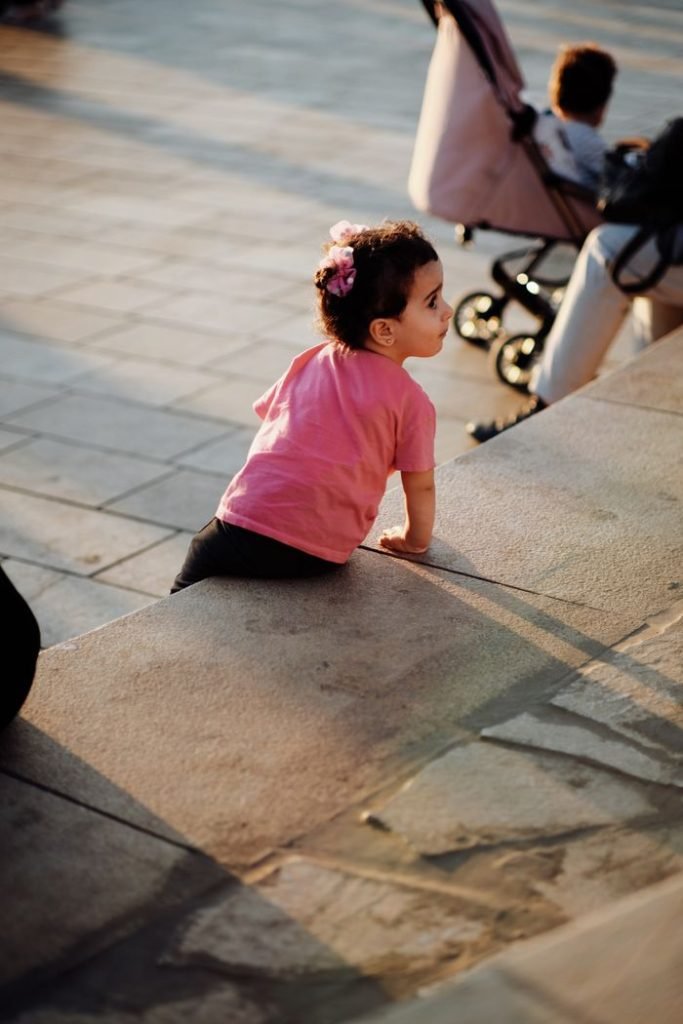 A young girl in a pink shirt exploring and climbing a city step, embodying innocence and curiosity.