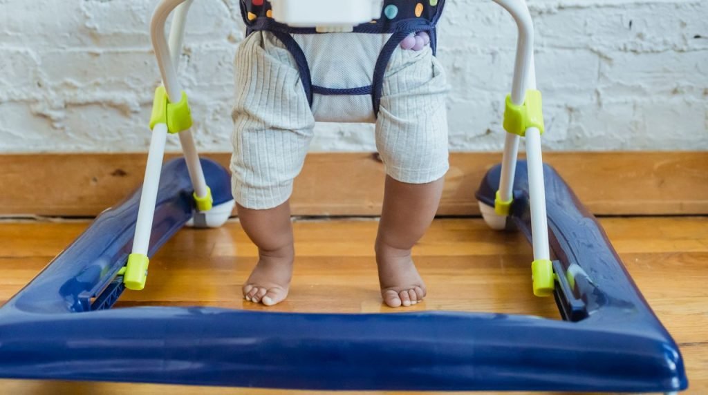 Unrecognizable barefoot African American toddler in casual clothes sitting in baby walker near white wall in light room at home