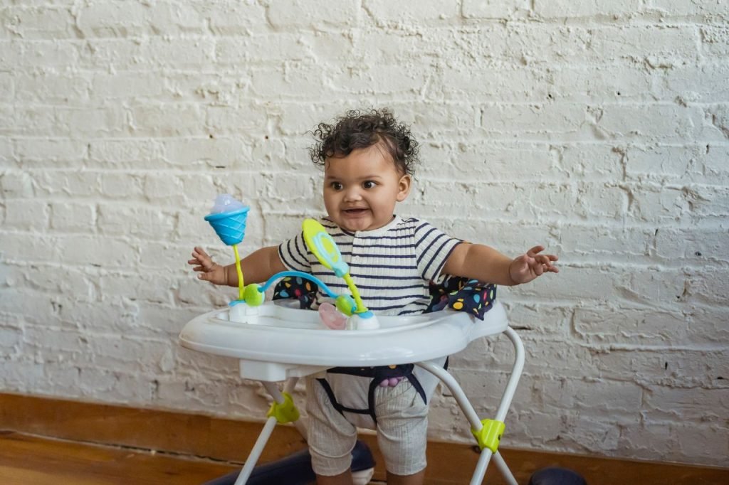Funny African American toddler with spread arms sitting in baby walkers with toys near white wall in light room at home