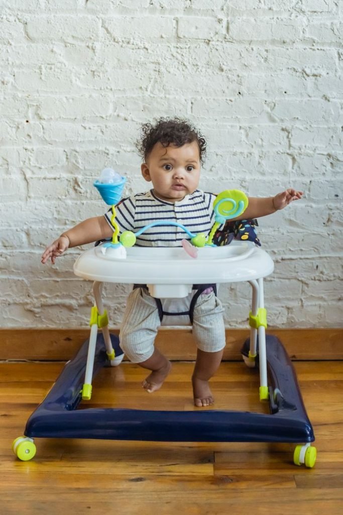 Full body of adorable barefoot African American baby in casual wear looking away while sitting in walker with toys near wall