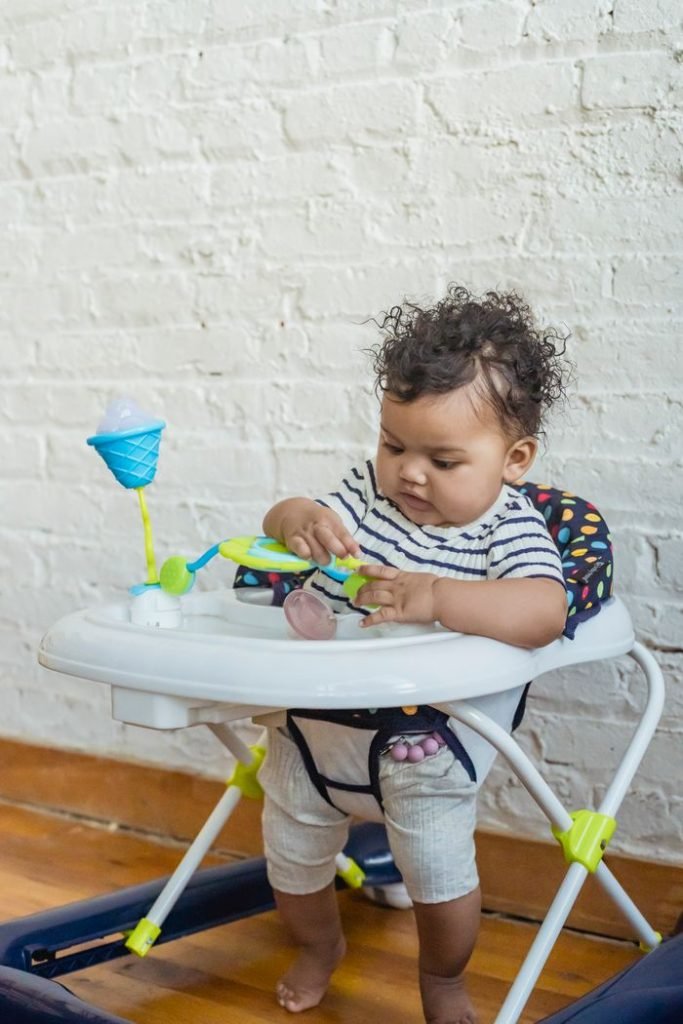 Curious little barefoot African American baby sitting in walker and playing with toy near white wall in room at home
